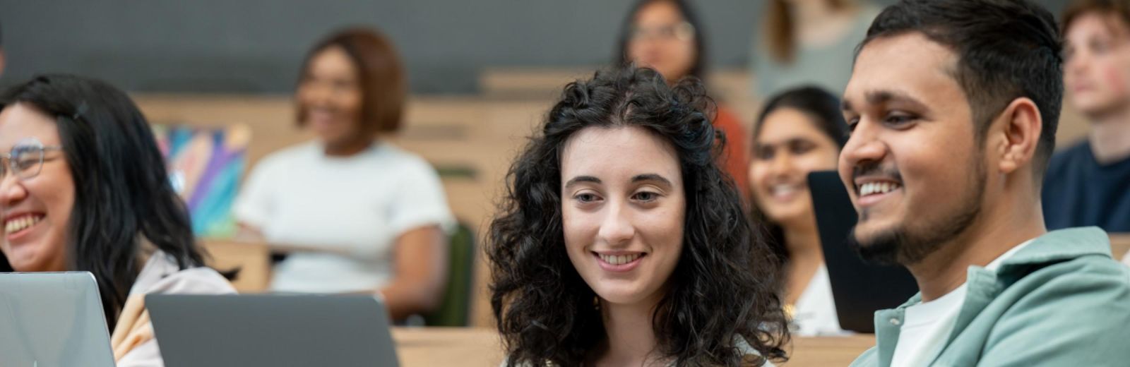 Many students are in a lecture theatre. Two students are in the foreground smiling and looking at a laptop.