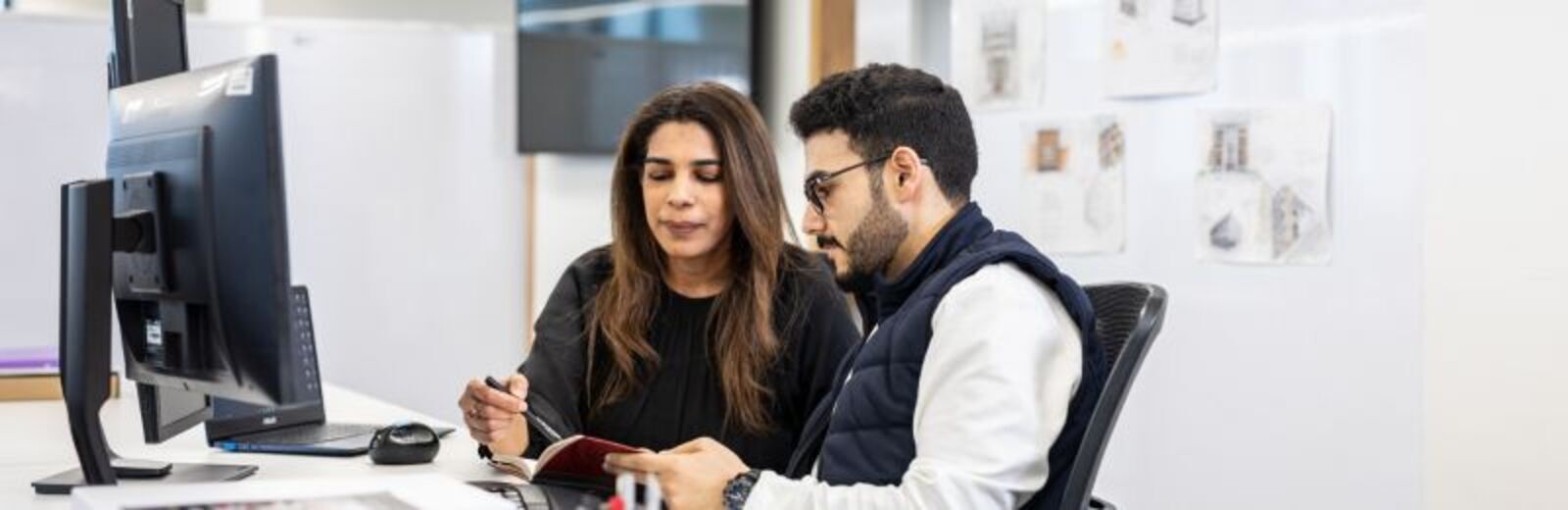 A student and their professor looking at a computer screen, with engineering plans stuck to the wall behind the.