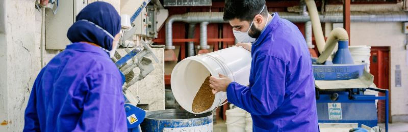 Two students working in a civil engineering lab, dressed in lab coats and PPE. One is pouring red cement power in to a metal drum.