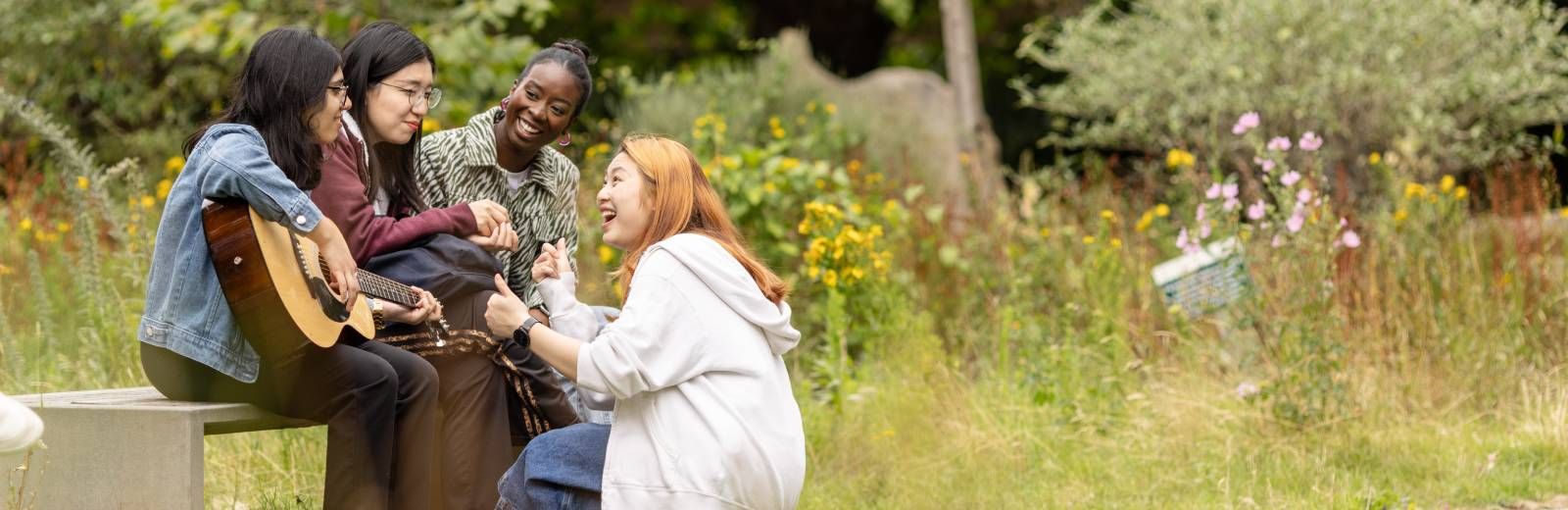 Four students together in the Sustainable Garden on campus. They are surrounded by wildflowers, one student holds a guitar in her hands whilst another crouches in front of her showing her how to play. The others watch on happily.