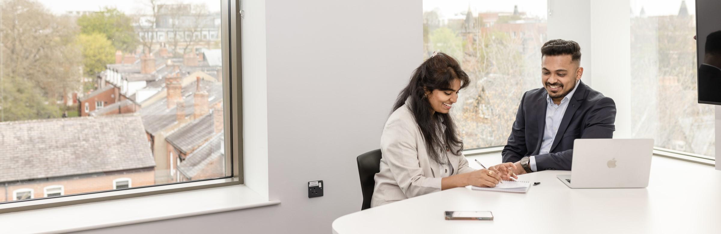 Two students sit together in a modern room with lots of windows. They are looking at a laptop screen.